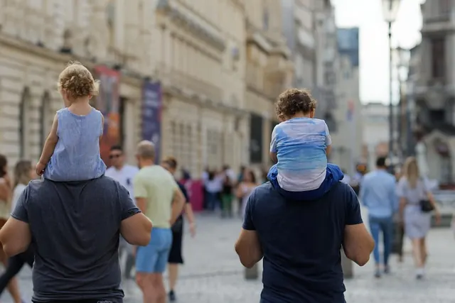 Two fathers walking through a city street with their children on their shoulders