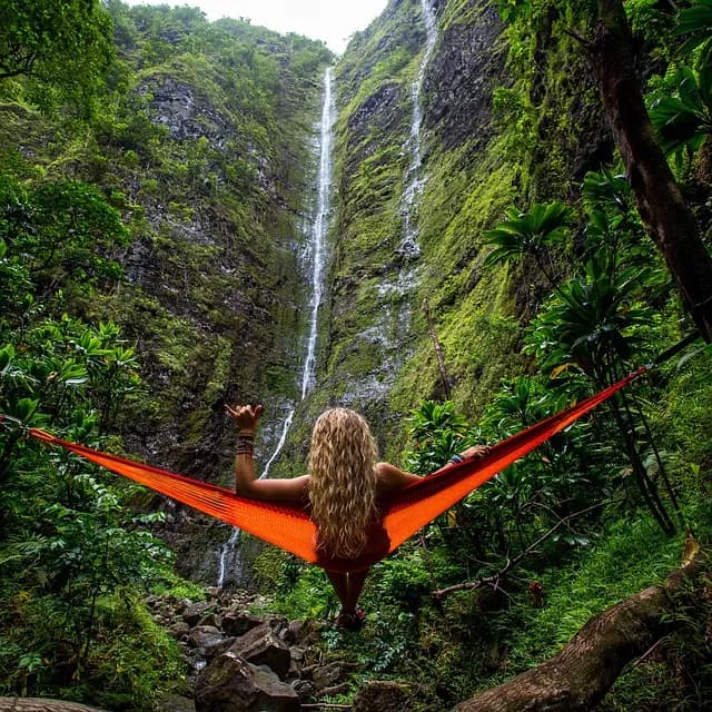 A person resting in a hammock facing a tall waterfall in dense green forest