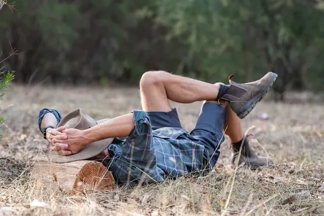 A man lying back in dry grass with his hat over his face, boots crossed